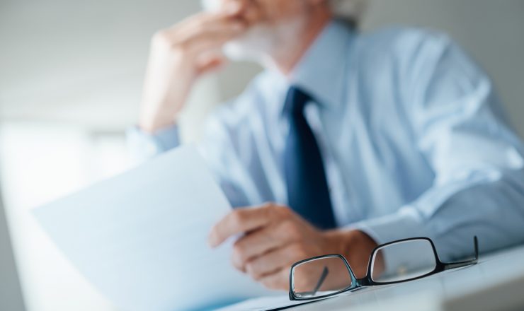 Pensive businessman with hand on chin looking away and holding a document, selective focus, glasses on foreground