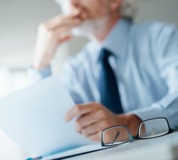 Pensive businessman with hand on chin looking away and holding a document, selective focus, glasses on foreground